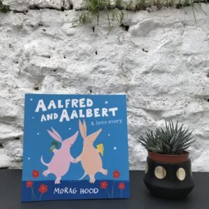 On a black table, a copy of 'Aalfred and Aalbert' by Morag Hood stands on a black table, next to a potted plant.  Behind them, a stone, white wall obscures the background.