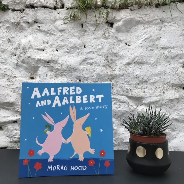 On a black table, a copy of 'Aalfred and Aalbert' by Morag Hood stands on a black table, next to a potted plant.  Behind them, a stone, white wall obscures the background.