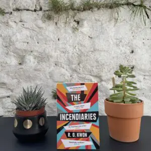 On a black table, between two potted plants, a copy of the book is standing, with a white, stone wall in the background.