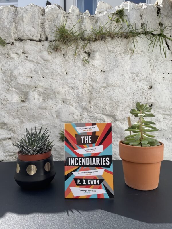 On a black table, between two potted plants, a copy of the book is standing, with a white, stone wall in the background.
