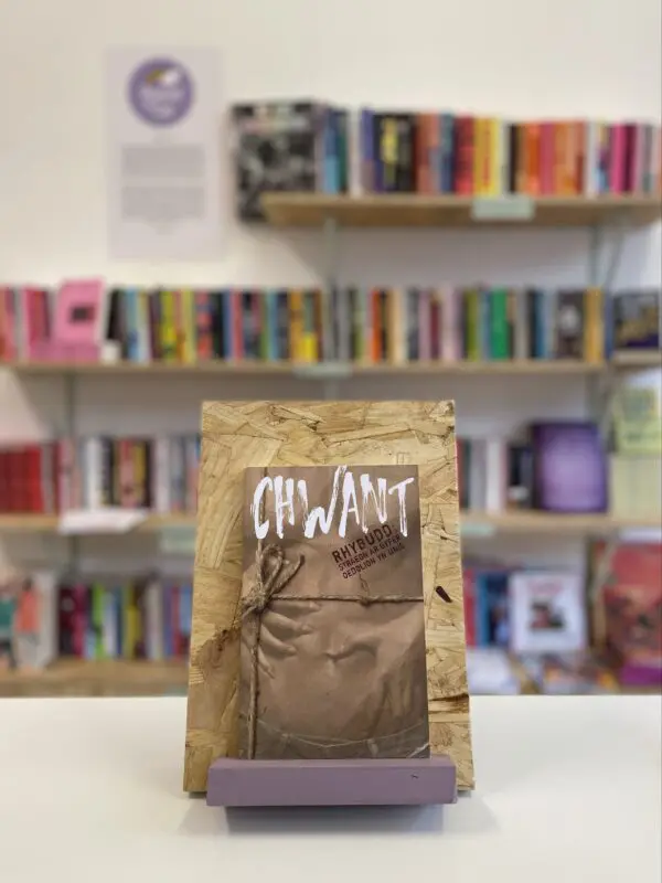 Cymraeg: Copi o 'Chwant' yn sefyll ar stondin llyfrau, tu blaen silffoedd o lyfrau yn y cefndir. | English: A copy of 'Chwant' sits on a stand in front of multiple shelves of other books.