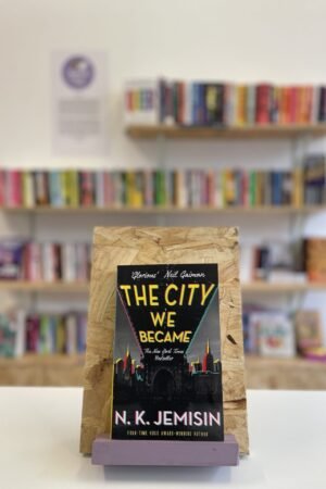 Cymraeg: Copi o 'The City We Became' yn sefyll ar stondin llyfrau, tu blaen silffoedd o lyfrau yn y cefndir. | English: A copy of 'The City We Became' sits on a stand in front of multiple shelves of other books.