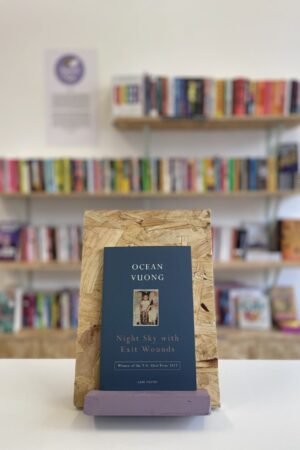 Cymraeg: Copi o 'Night Sky with Exit Wounds' yn sefyll ar stondin llyfrau, tu blaen silffoedd o lyfrau yn y cefndir. | English: A copy of 'Night Sky with Exit Wounds' sits on a stand in front of multiple shelves of other books.