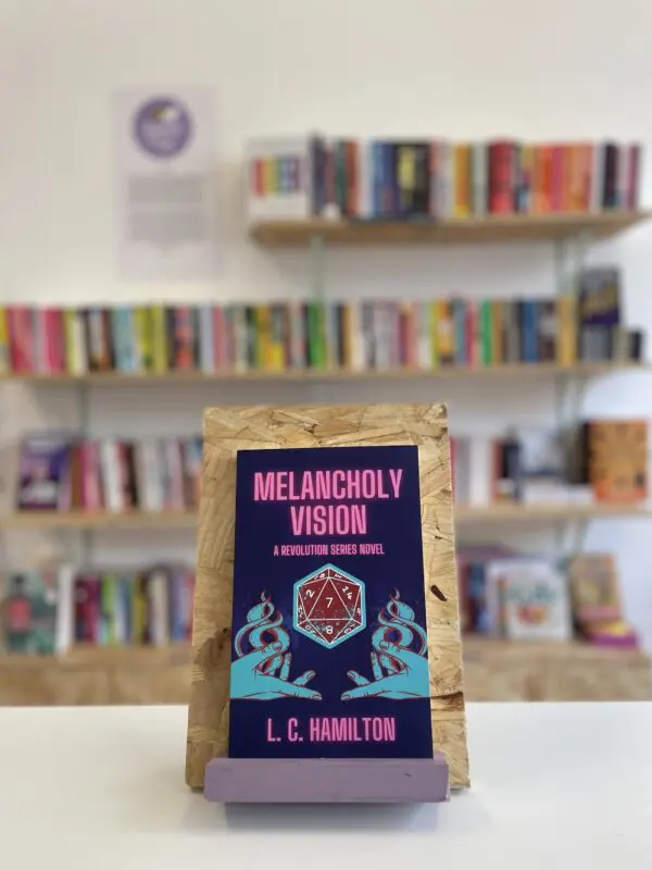 Cymraeg: Copi o 'Melancholy Vision' yn sefyll ar stondin llyfrau, tu blaen silffoedd o lyfrau yn y cefndir. | English: A copy of 'Melancholy Vision' sits on a stand in front of multiple shelves of other books.