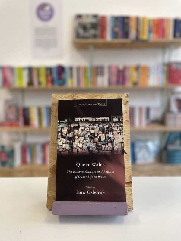 A copy of 'Queer Wales' sits on a stand in front of multiple shelves of other books.