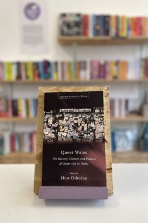 A copy of 'Queer Wales' sits on a stand in front of multiple shelves of other books.