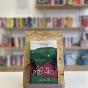Cymraeg: Copi o 'On the Red Hill' yn sefyll ar stondin llyfrau, tu blaen silffoedd o lyfrau yn y cefndir. | English: A copy of 'On the Red Hill' sits on a stand in front of multiple shelves of other books.