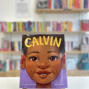 Cymraeg: Copi o 'Calvin' yn sefyll ar stondin llyfrau, tu blaen silffoedd o lyfrau yn y cefndir. | English: A copy of 'Calvin' sits on a stand in front of multiple shelves of other books.