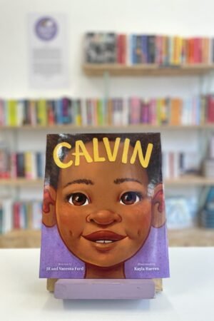 Cymraeg: Copi o 'Calvin' yn sefyll ar stondin llyfrau, tu blaen silffoedd o lyfrau yn y cefndir. | English: A copy of 'Calvin' sits on a stand in front of multiple shelves of other books.