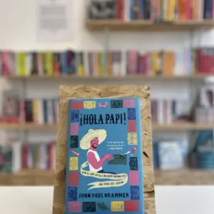 Cymraeg: Copi o 'Hola Papi!' yn sefyll ar stondin llyfrau, tu blaen silffoedd o lyfrau yn y cefndir. | English: A copy of 'Hola Papi!' sits on a stand in front of multiple shelves of other books.