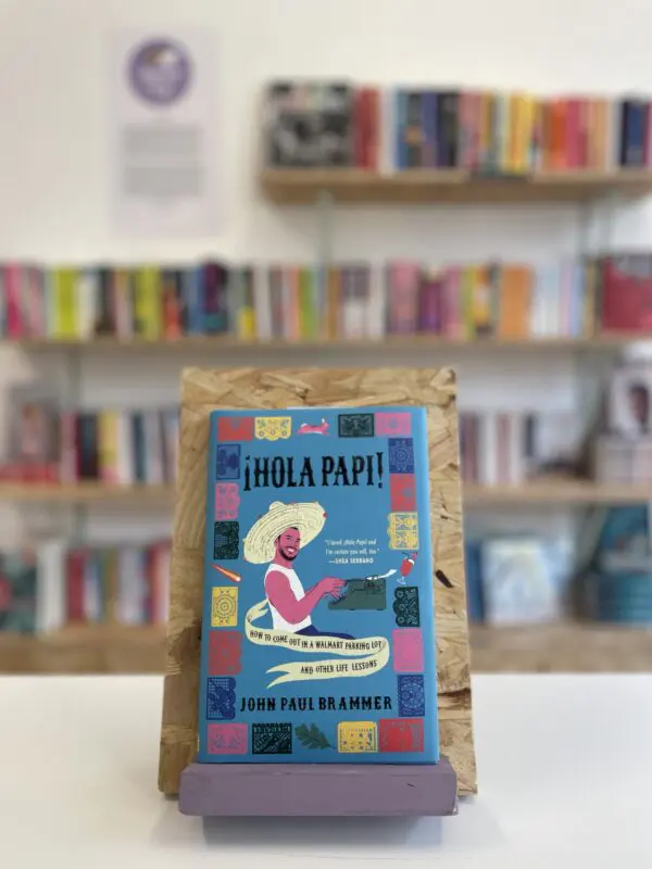 Cymraeg: Copi o 'Hola Papi!' yn sefyll ar stondin llyfrau, tu blaen silffoedd o lyfrau yn y cefndir. | English: A copy of 'Hola Papi!' sits on a stand in front of multiple shelves of other books.