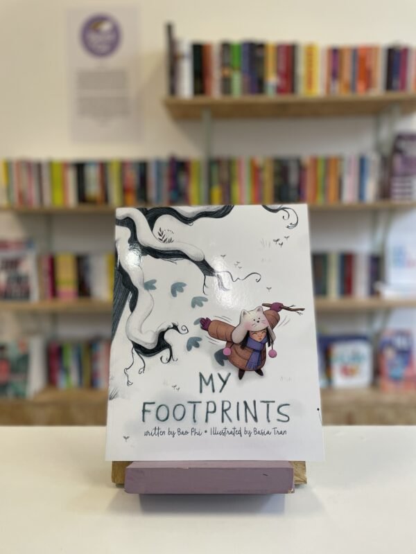 Cymraeg: Copi o 'My Footprints' yn sefyll ar stondin llyfrau, tu blaen silffoedd o lyfrau yn y cefndir. | English: A copy of 'My Footprints' sits on a stand in front of multiple shelves of other books.