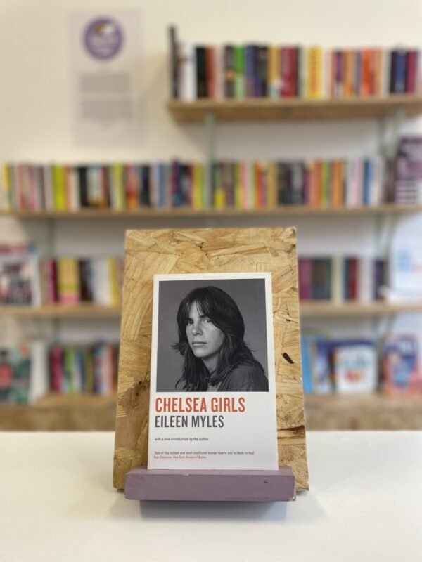 Cymraeg: Copi o 'Chelsea Girls' yn sefyll ar stondin llyfrau, tu blaen silffoedd o lyfrau yn y cefndir. | English: A copy of 'Chelsea Girls' sits on a stand in front of multiple shelves of other books.