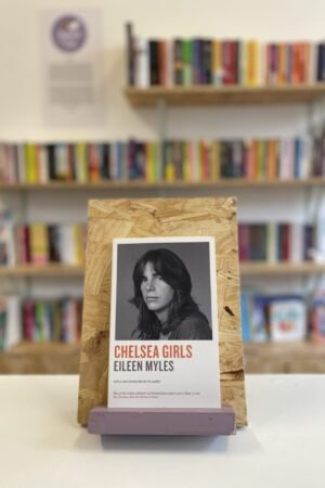 Cymraeg: Copi o 'Chelsea Girls' yn sefyll ar stondin llyfrau, tu blaen silffoedd o lyfrau yn y cefndir. | English: A copy of 'Chelsea Girls' sits on a stand in front of multiple shelves of other books.