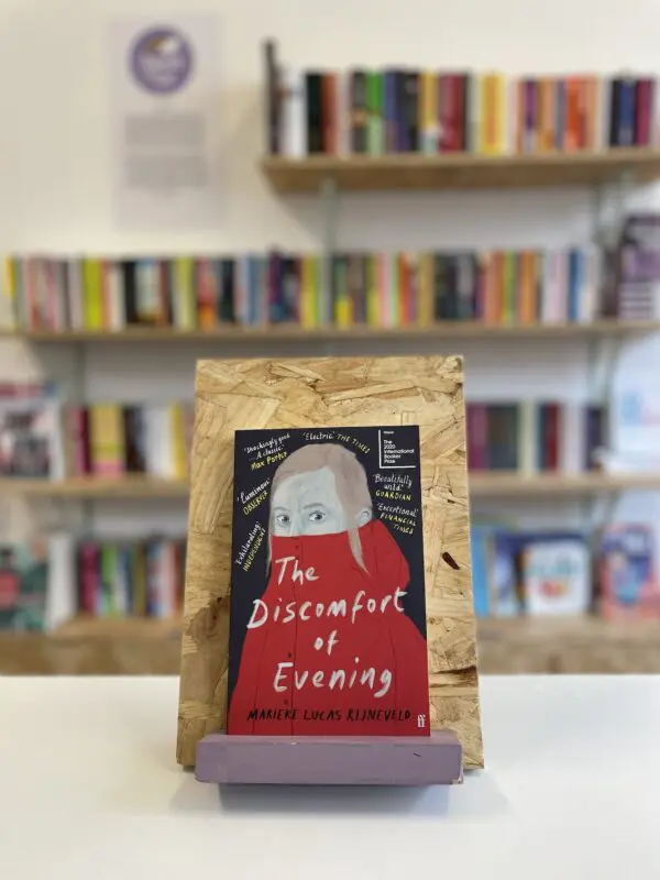 Cymraeg: Copi o 'The Discomfort of Evening' yn sefyll ar stondin llyfrau, tu blaen silffoedd o lyfrau yn y cefndir. | English: A copy of 'Deep Sniff' sits on a stand in front of multiple shelves of other books.