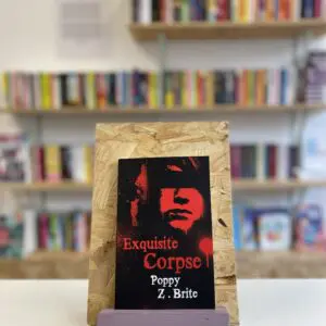 Cymraeg: Copi o 'Exquisite Corpse' yn sefyll ar stondin llyfrau, tu blaen silffoedd o lyfrau yn y cefndir. | English: A copy of 'Exquisite Corpse' sits on a stand in front of multiple shelves of other books.