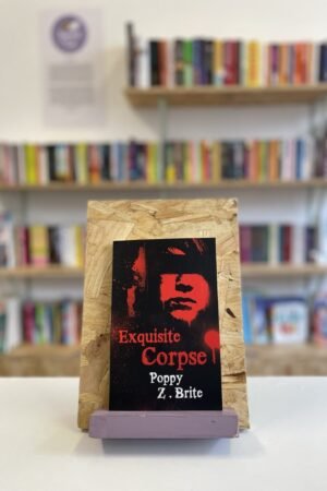 Cymraeg: Copi o 'Exquisite Corpse' yn sefyll ar stondin llyfrau, tu blaen silffoedd o lyfrau yn y cefndir. | English: A copy of 'Exquisite Corpse' sits on a stand in front of multiple shelves of other books.