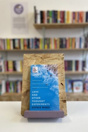 Cymraeg: Copi o 'Love and Other Thought Experiments' yn sefyll ar stondin llyfrau, tu blaen silffoedd o lyfrau yn y cefndir. | English: A copy of 'Love and Other Thought Experiments' sits on a stand in front of multiple shelves of other books.
