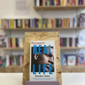 Cymraeg: Copi o 'Real Life' yn sefyll ar stondin llyfrau, tu blaen silffoedd o lyfrau yn y cefndir. | English: A copy of 'Real Life' sits on a stand in front of multiple shelves of other books.