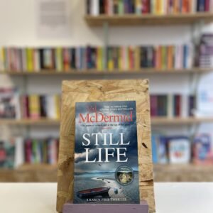 Cymraeg: Copi o 'Still Life' yn sefyll ar stondin llyfrau, tu blaen silffoedd o lyfrau yn y cefndir. | English: A copy of 'Still Life' sits on a stand in front of multiple shelves of other books.