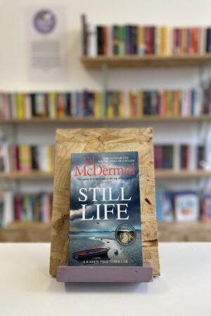 Cymraeg: Copi o 'Still Life' yn sefyll ar stondin llyfrau, tu blaen silffoedd o lyfrau yn y cefndir. | English: A copy of 'Still Life' sits on a stand in front of multiple shelves of other books.