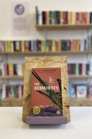 Cymraeg: Copi o 'The Remainder' yn sefyll ar stondin llyfrau, tu blaen silffoedd o lyfrau yn y cefndir. | English: A copy of 'The Remainder' sits on a stand in front of multiple shelves of other books.