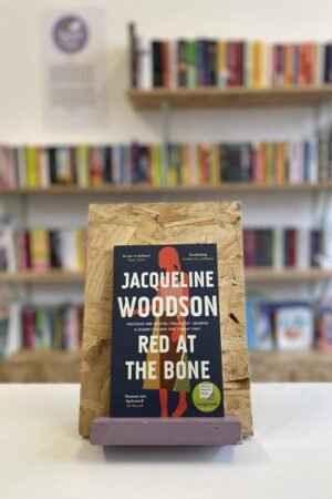 Cymraeg: Copi o 'Red at the Bone' yn sefyll ar stondin llyfrau, tu blaen silffoedd o lyfrau yn y cefndir. | English: A copy of 'Red at the Bone' sits on a stand in front of multiple shelves of other books.