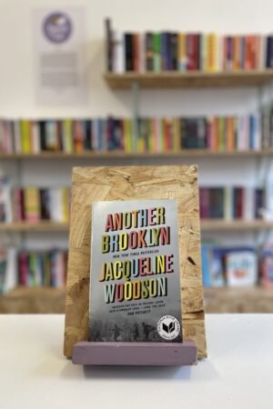 Cymraeg: Copi o 'Another Brooklyn' yn sefyll ar stondin llyfrau, tu blaen silffoedd o lyfrau yn y cefndir. | English: A copy of 'Another Brooklyn' sits on a stand in front of multiple shelves of other books.