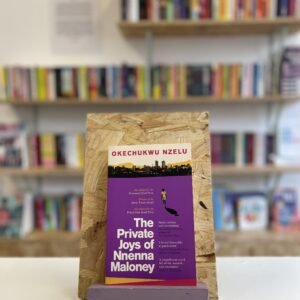 A copy of 'The Private Joys of Nnenna Maloney' sits on a stand in front of multiple shelves of other books.