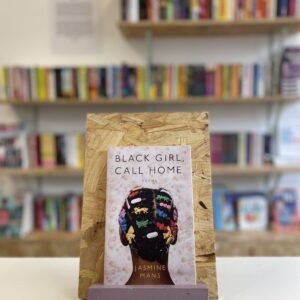 Cymraeg: Copi o 'Black Girl, Call Home' yn sefyll ar stondin llyfrau, tu blaen silffoedd o lyfrau yn y cefndir. | English: A copy of 'Black Girl, Call Home' sits on a stand in front of multiple shelves of other books.