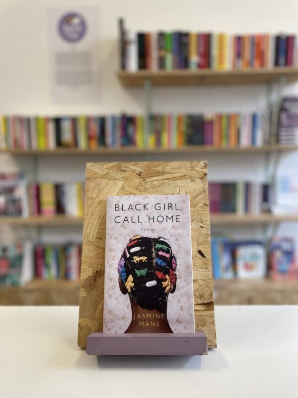 Cymraeg: Copi o 'Black Girl, Call Home' yn sefyll ar stondin llyfrau, tu blaen silffoedd o lyfrau yn y cefndir. | English: A copy of 'Black Girl, Call Home' sits on a stand in front of multiple shelves of other books.