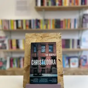 Cymraeg: Copi o 'Christodora' yn sefyll ar stondin llyfrau, tu blaen silffoedd o lyfrau yn y cefndir. | English: A copy of 'Christodora' sits on a stand in front of multiple shelves of other books.