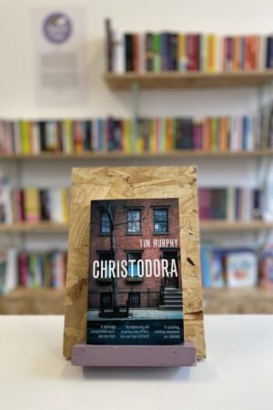 Cymraeg: Copi o 'Christodora' yn sefyll ar stondin llyfrau, tu blaen silffoedd o lyfrau yn y cefndir. | English: A copy of 'Christodora' sits on a stand in front of multiple shelves of other books.