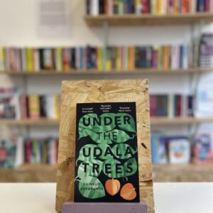 Cymraeg: Copi o 'Under the Udala Trees' yn sefyll ar stondin llyfrau, tu blaen silffoedd o lyfrau yn y cefndir. | English: A copy of 'Under the Udala Trees' sits on a stand in front of multiple shelves of other books.