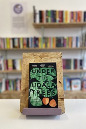 Cymraeg: Copi o 'Under the Udala Trees' yn sefyll ar stondin llyfrau, tu blaen silffoedd o lyfrau yn y cefndir. | English: A copy of 'Under the Udala Trees' sits on a stand in front of multiple shelves of other books.
