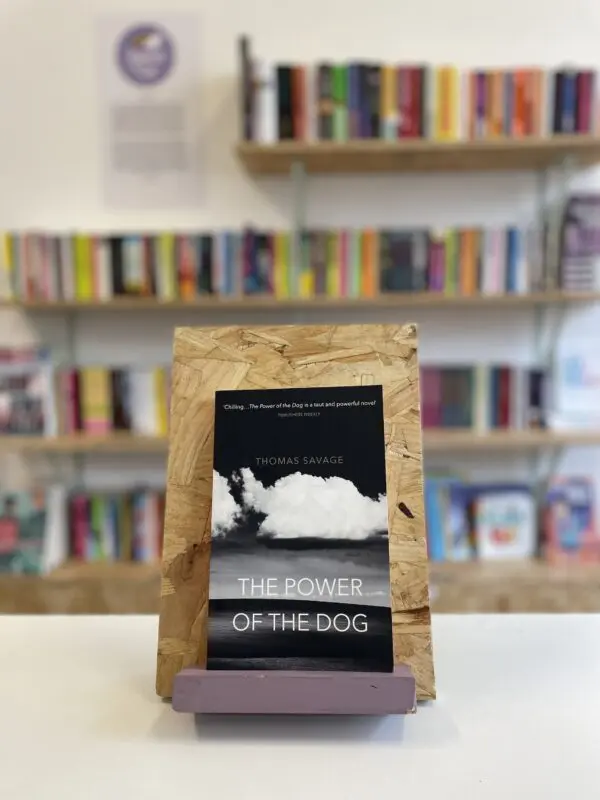 Cymraeg: Copi o 'The Power of the Dog' yn sefyll ar stondin llyfrau, tu blaen silffoedd o lyfrau yn y cefndir. | English: A copy of 'The Power of the Dog' sits on a stand in front of multiple shelves of other books.
