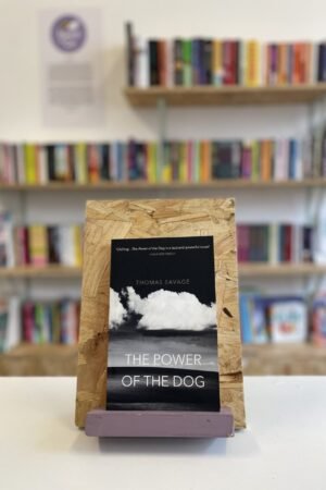 Cymraeg: Copi o 'The Power of the Dog' yn sefyll ar stondin llyfrau, tu blaen silffoedd o lyfrau yn y cefndir. | English: A copy of 'The Power of the Dog' sits on a stand in front of multiple shelves of other books.