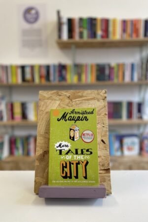 Cymraeg: Copi o 'More Tales of the City' yn sefyll ar stondin llyfrau, tu blaen silffoedd o lyfrau yn y cefndir. | English: A copy of 'More Tales of the City' sits on a stand in front of multiple shelves of other books.