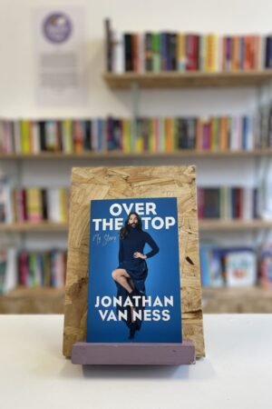 Cymraeg: Copi o 'Over the top' yn sefyll ar stondin llyfrau, tu blaen silffoedd o lyfrau yn y cefndir. | English: A copy of 'Over the Top' sits on a stand in front of multiple shelves of other books.