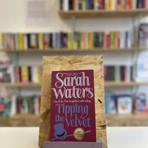 Cymraeg: Copi o 'Tipping the Velvet' yn sefyll ar stondin llyfrau, tu blaen silffoedd o lyfrau yn y cefndir. | English: A copy of 'Tipping the Velvet' sits on a stand in front of multiple shelves of other books.