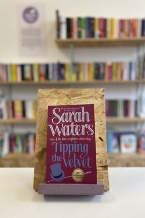 Cymraeg: Copi o 'Tipping the Velvet' yn sefyll ar stondin llyfrau, tu blaen silffoedd o lyfrau yn y cefndir. | English: A copy of 'Tipping the Velvet' sits on a stand in front of multiple shelves of other books.