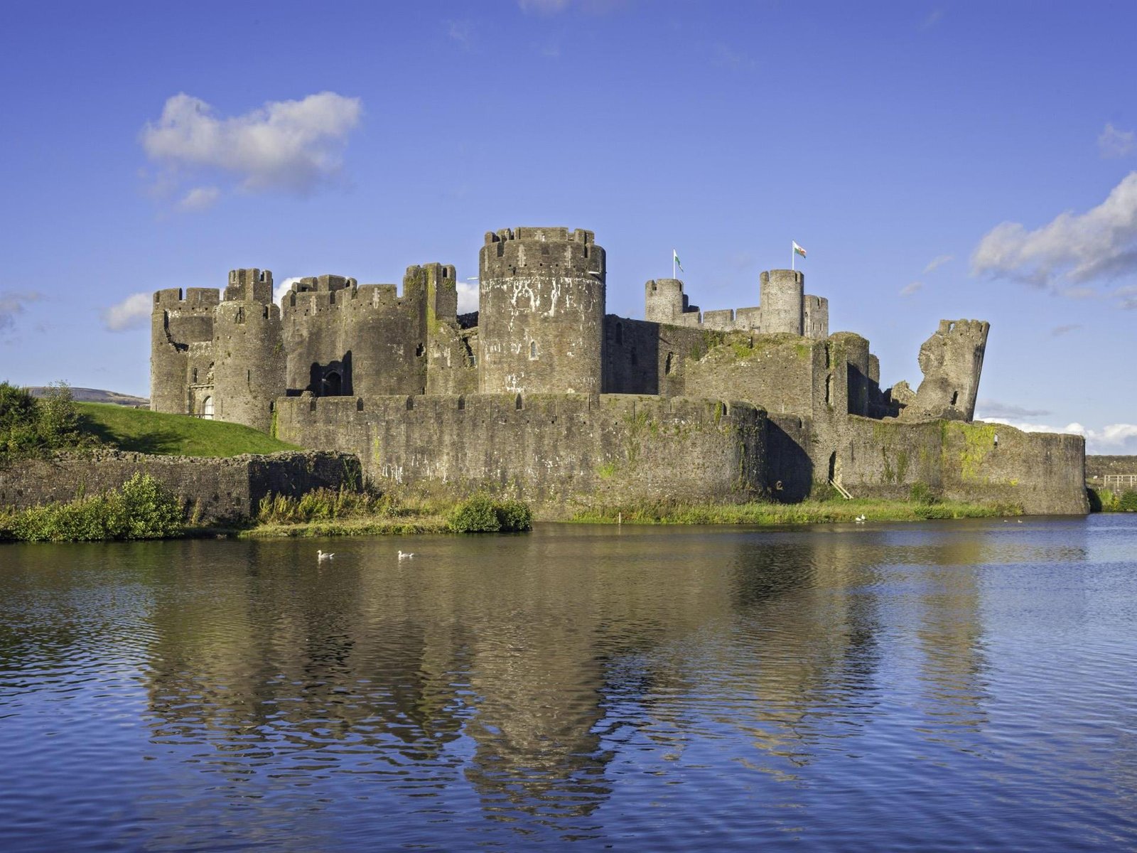 In front of Caerphilly Castle. there is a body of water and above it, a clear blue sky.