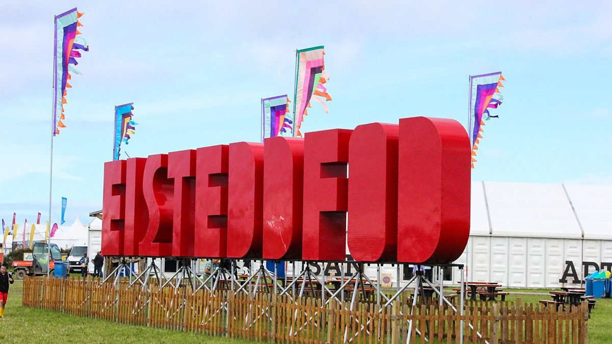 Cerflun o'r gair 'Eisteddfod' mewn coch ar lwyfan mewn cae gwyrdd. Yn y cefndir, mae yna faneri, adeilad perfformio wyn ac awyr glir.