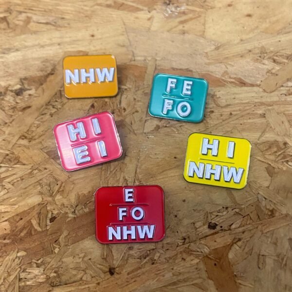 Cymraeg: Ar arwyneb pren, mae yna pump pin rhagenwau mewn lliwiau gwahanol: ‘E/Fe/Fo’ (coch/red) ‘Hi/Ei’ (pinc/pink) ‘Nhw’ (oren) ‘Fe/Fo’ (gwyrdd) ‘Hi/Nhw’ (melyn/yellow) English: The aforementioned Welsh language pronouns pins are sitting on a wooden surface.
