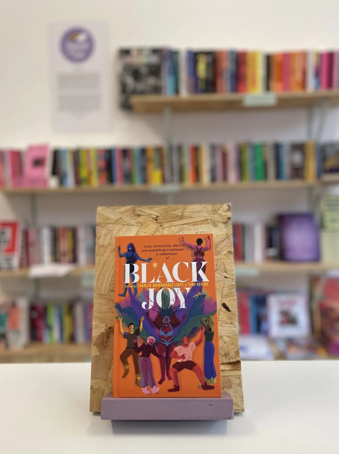 Cymraeg: Copi o 'Black Joy' yn sefyll ar stondin llyfrau, tu blaen silffoedd o lyfrau yn y cefndir. English: A copy of 'Black Joy' sits on a stand in front of multiple shelves of other books.