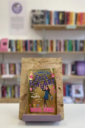 A copy of 'The Amazing Edie Eckhart' sits on a stand in front of multiple shelves of other books.