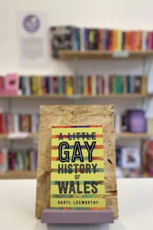 A copy of 'A Little Gay History of Wales' sits on a stand in front of multiple shelves of other books.