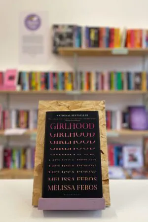A copy of 'Girlhood' sits on a stand in front of multiple shelves of other books.