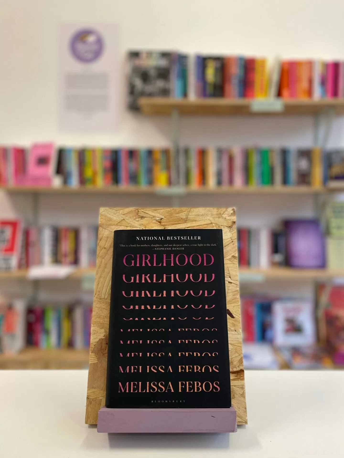 A copy of 'Girlhood' sits on a stand in front of multiple shelves of other books.