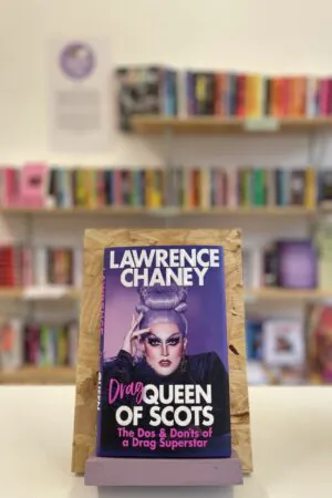 Cymraeg: Copi o 'Drag Queen of Scots' yn sefyll ar stondin llyfrau, tu blaen silffoedd o lyfrau yn y cefndir. | English: A copy of 'Drag Queen of Scots' sits on a stand in front of multiple shelves of other books.